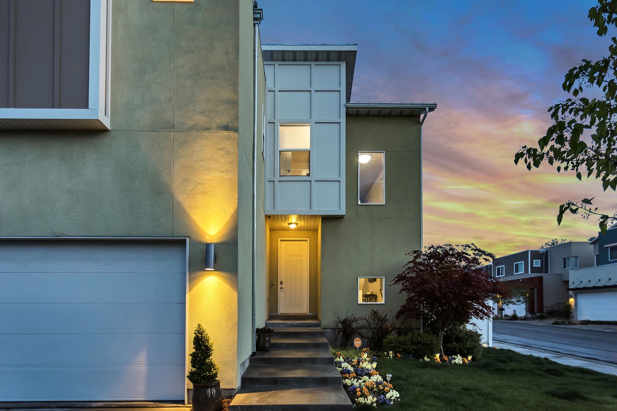 A house entrance with lit wall lamps and steps under a pink dusk sky